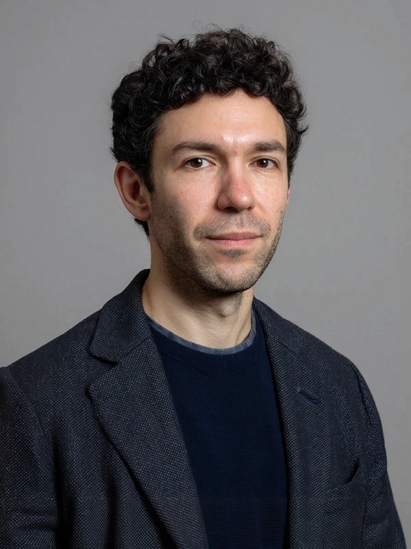 A young man with curly dark hair, wearing a black t-shirt and blazer, poses against a plain background.