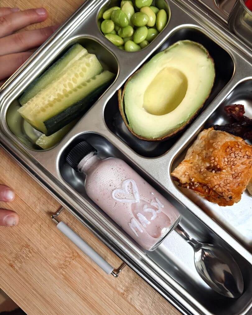 Balanced kids lunchbox with avocado half, cucumber sticks, edamame, sesame pastry and berry smoothie.