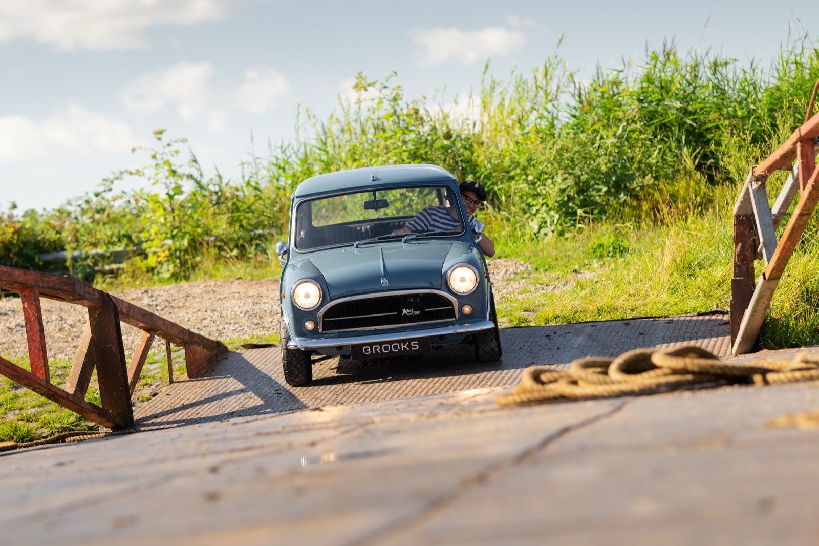 A classic car drives over a small wooden bridge surrounded by lush green trees and a clear blue sky.