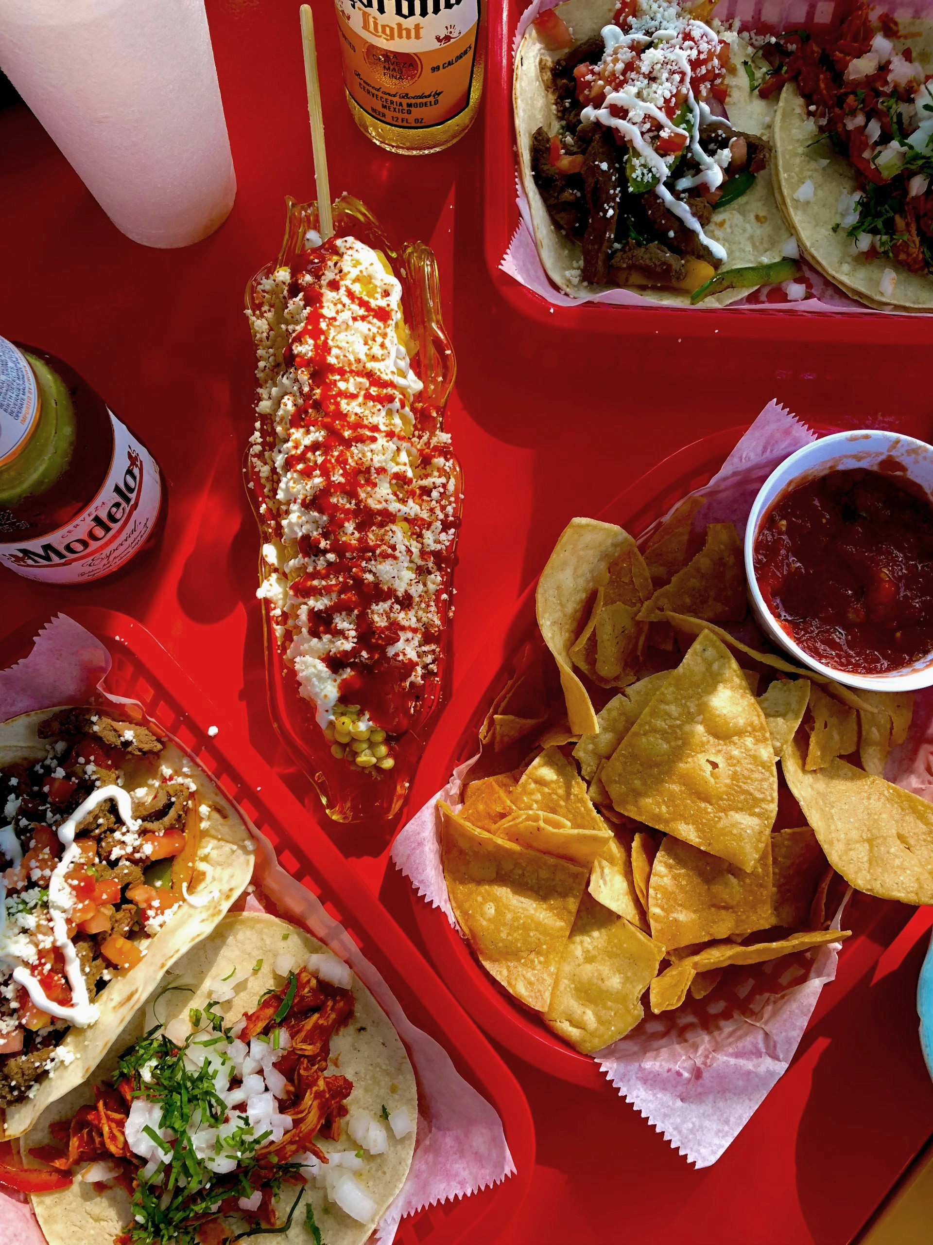 Top-down view of a bright red table covered with tacos, a dressed Mexican street corn, chips, salsa, and beer.