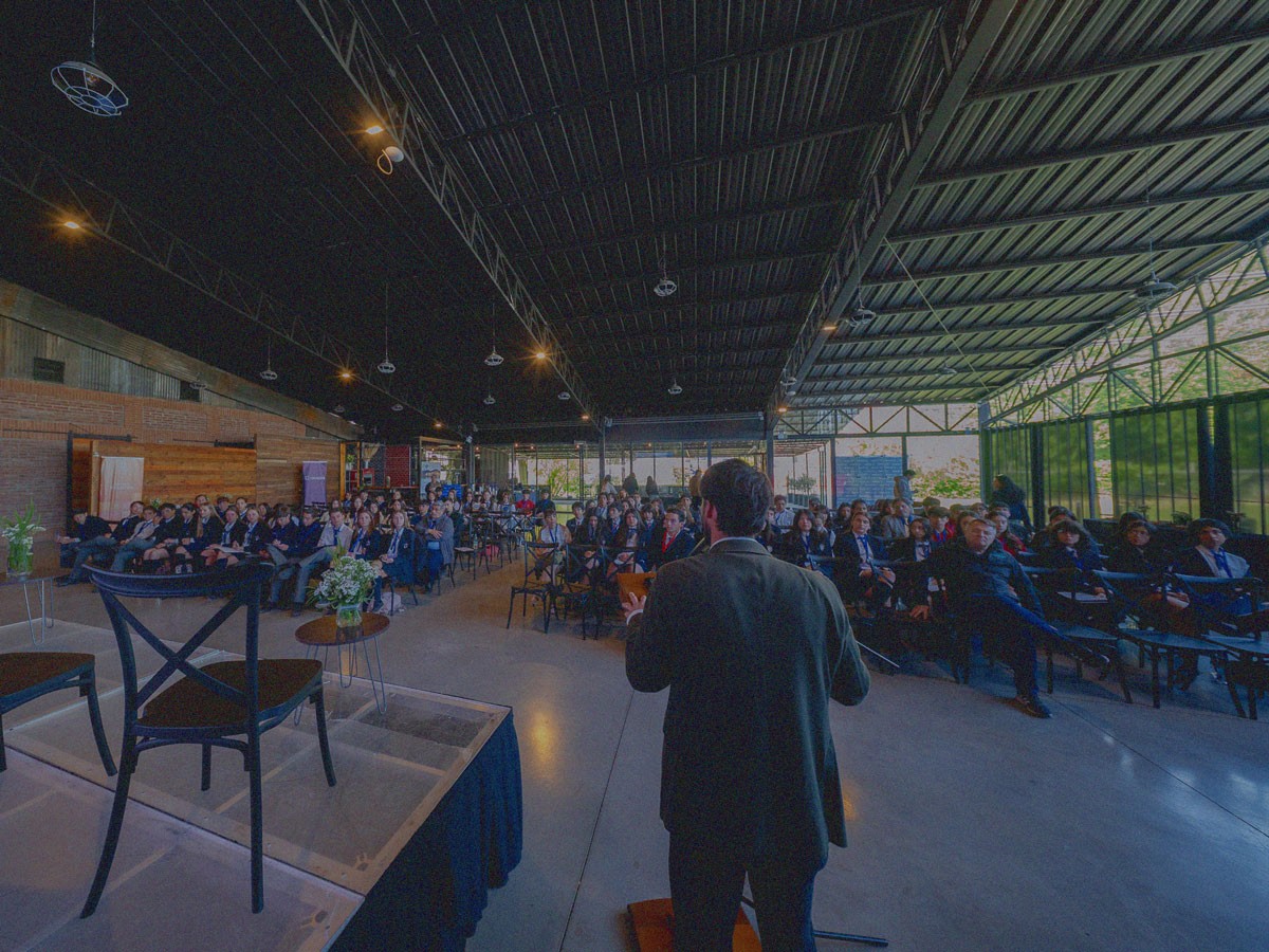 Persona de espaldas expone frente a espacio lleno de estudiantes escolares que escuchan sentados en un evento de IdeaPaís en la Región de O’Higgins.