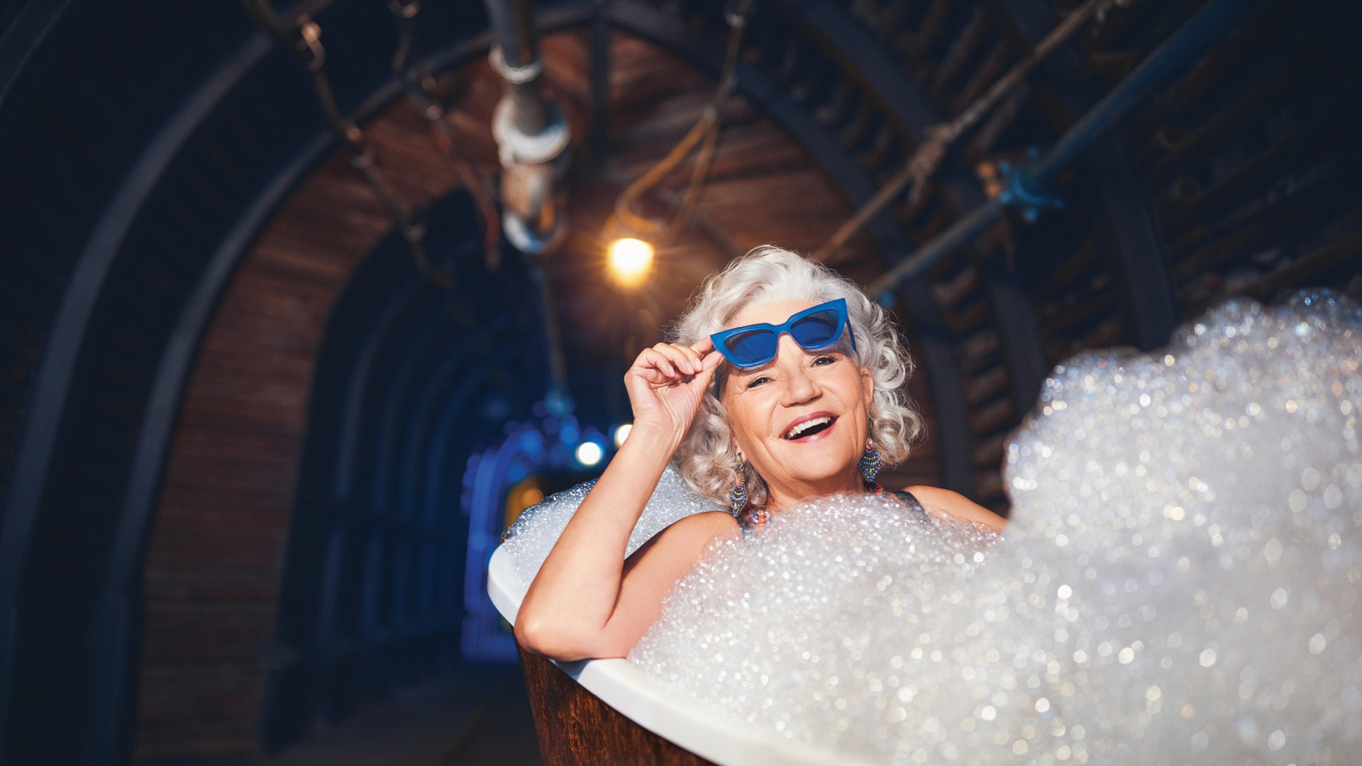Seniorin mit Sonnenbrille liegt lächelnd in einer Badewanne mit viel Schaum in einem beleuchtetem Grubenschacht