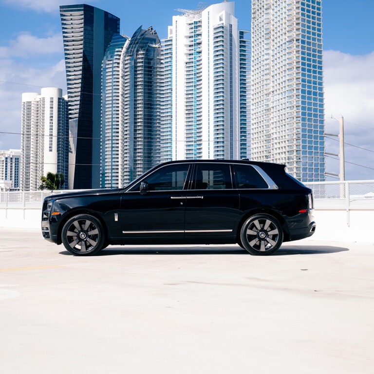 Side profile of a black Rolls-Royce Cullinan with city buildings in the background.
