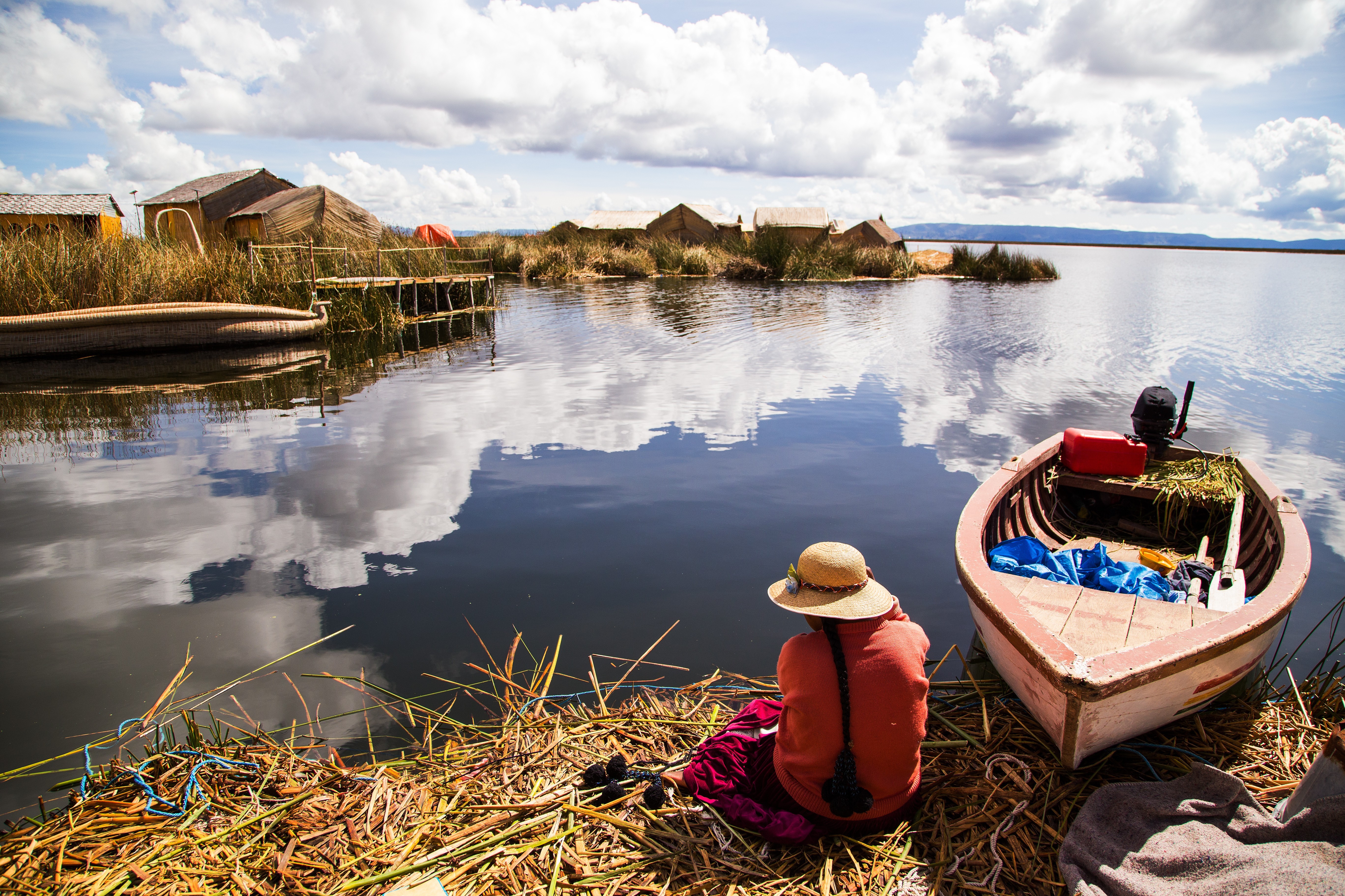 A woman in traditional clothing sits by a reed-lined shore beside a small boat, gazing across the still waters of Lake Titicaca toward floating reed islands under a sky filled with clouds.