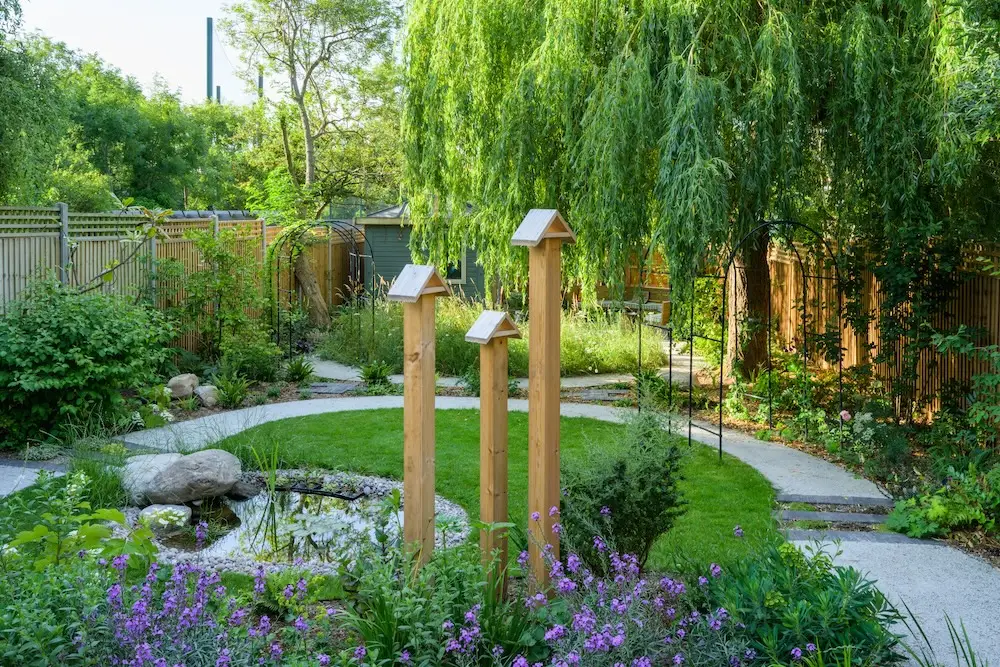 A serene garden path curves through lush greenery, featuring trees, flowers, and decorative wooden posts.