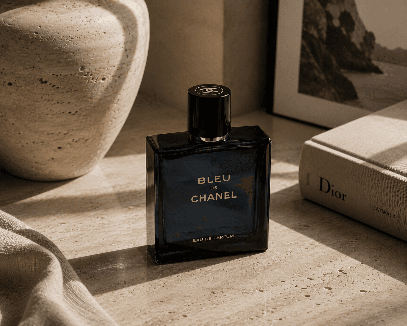 A square dark blue glass bottle of Bleu de Chanel Eau de Parfum with gold lettering and a black cylindrical cap stamped with the interlocking double-C logo. The perfume sits on a sunlit beige travertine surface next to a textured ceramic vase, a hardcover Dior book, a framed black-and-white photograph, and soft draped linen fabric.