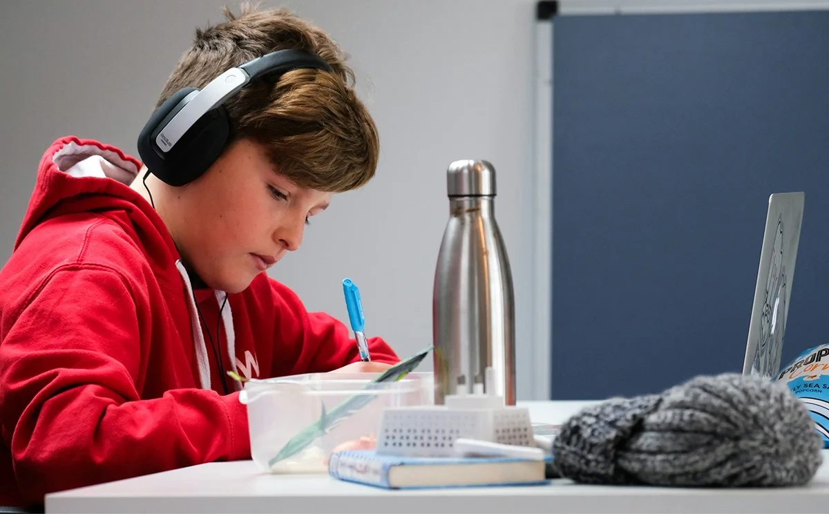A boy wearing headphones and writing beside a laptop.