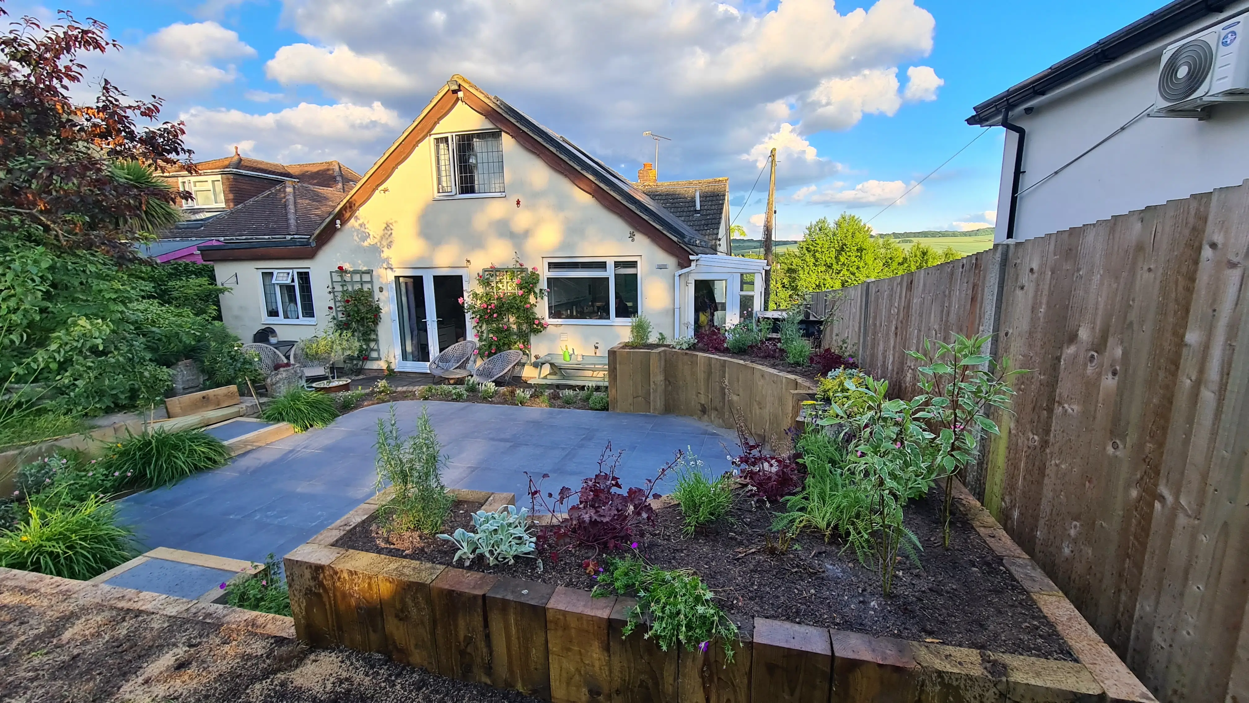 A sunny garden with plants, a patio area, and a house in the background, enclosed by wooden fencing.