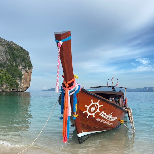 A colorful wooden boat with decorative ribbons is docked on a sandy beach, with a rocky cliff and calm sea in the background.