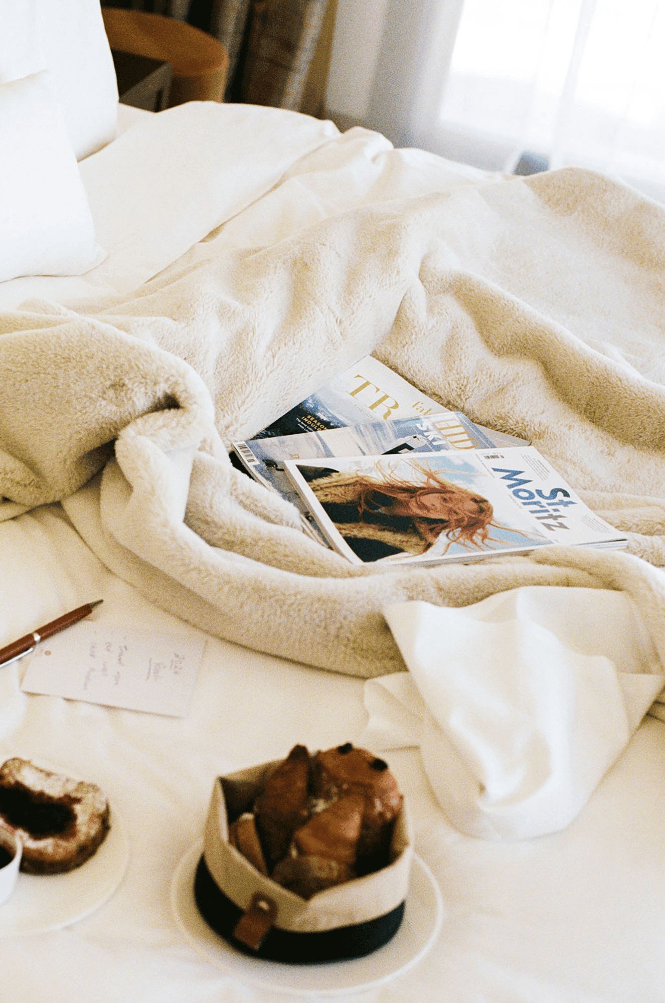Ski and travel magazines and a handwritten note rest on a fluffy white blanket, with pastries nearby