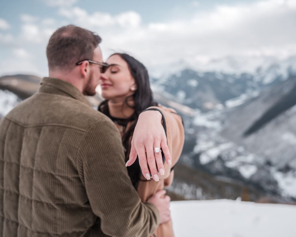 Couple embracing immediately after an Aspen mountain proposal, newly engaged woman extending her ring hand toward the camera with snow-covered peaks behind them