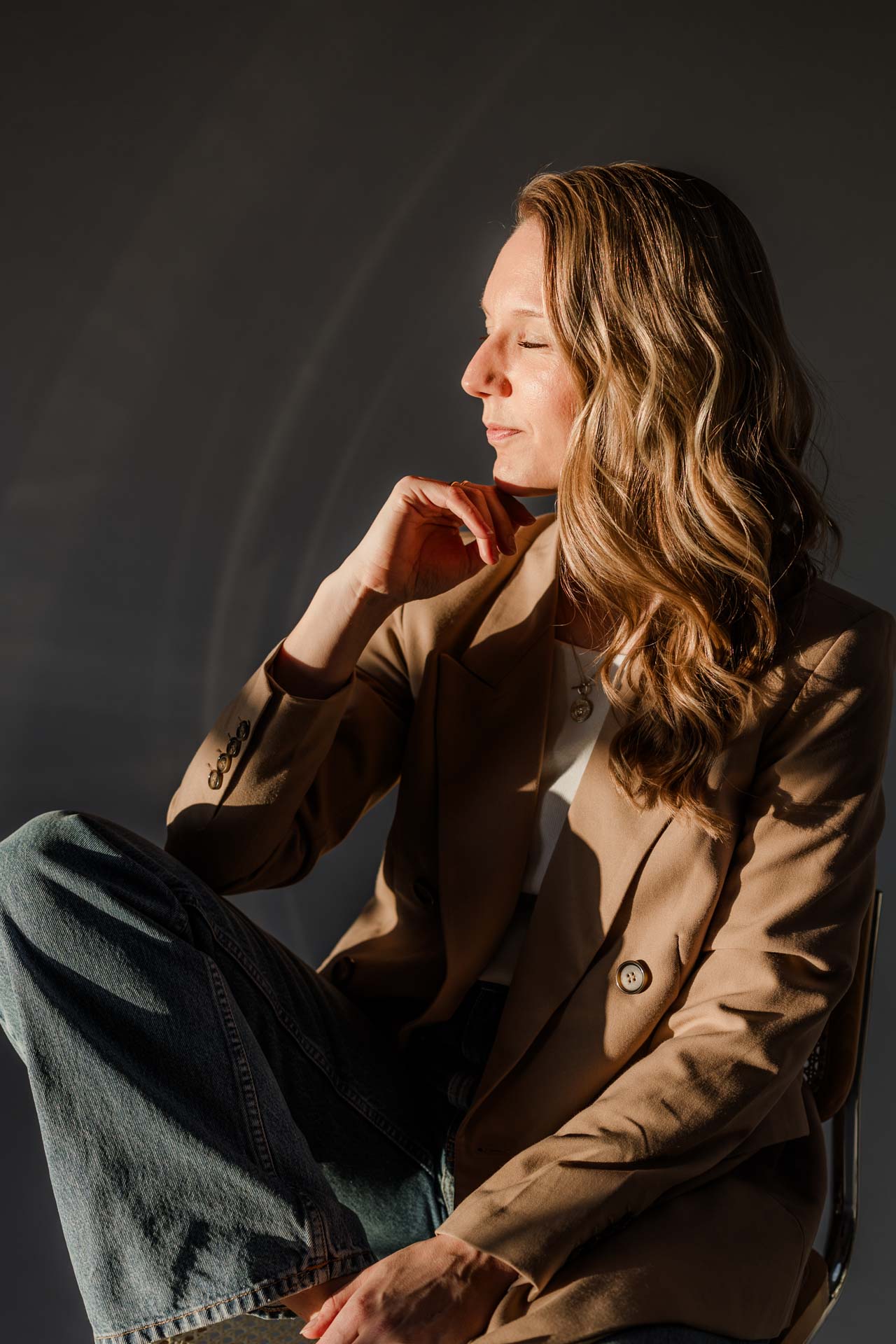 Die Gründerin aus Straubing, Veronika, von OHNORD studio sitzt von der Sonne angestahlt, in sich ruhend, mit geschlossenen Augen auf einem Stuhl. Ihr Outfit aus beigem Blazer und Jeans strahlt genau die Ruhe aus, die sie als Gründerin brauch.