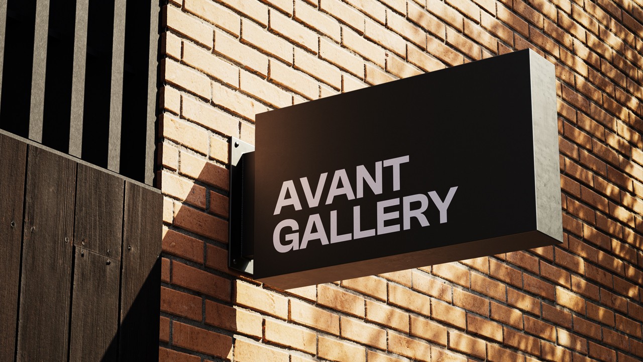 Black rectangular projecting sign mounted to a brick exterior wall, displaying “AVANT GALLERY” in light grey uppercase against the shadows and texture of the bricks.
