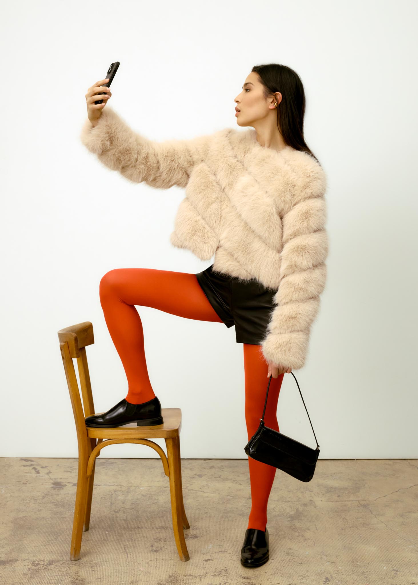 Femme prenant un selfie, un pied posé sur une chaise, portant une veste claire et des collants rouges