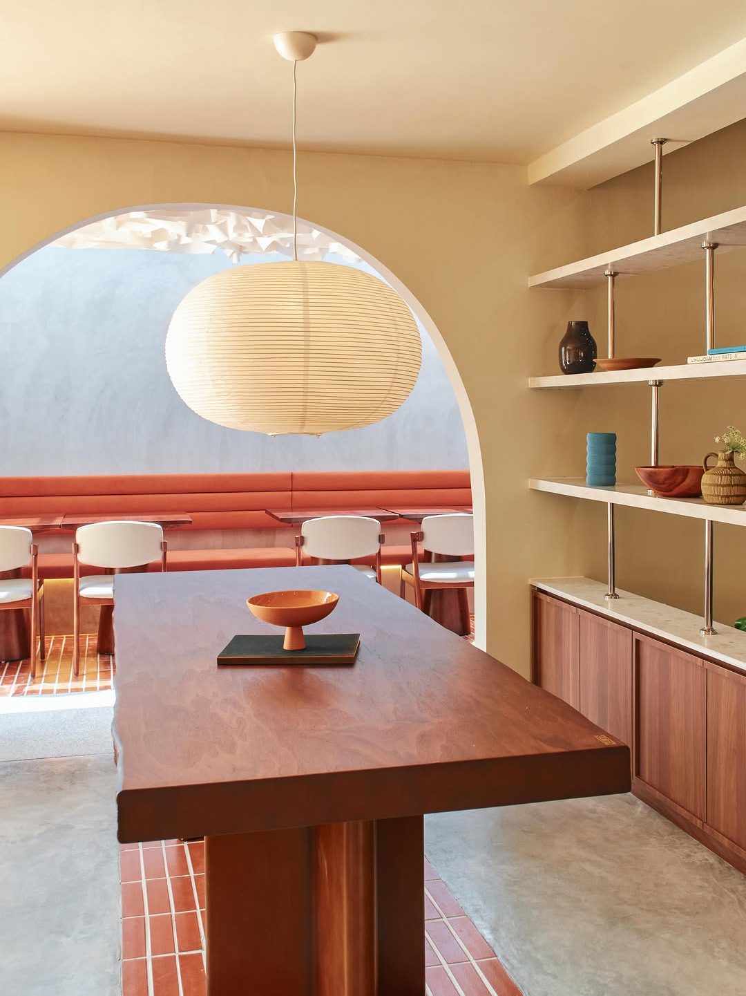 Interior view of a modern dining area featuring a large wooden table, minimalist decor, and a round paper lantern hanging overhead. Shelves with decorative items line the wall, and a red brick-patterned floor complements the space.