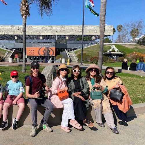 A group of six people sitting outdoors near the La Brea Tar Pits and Museum entrance on a sunny day.