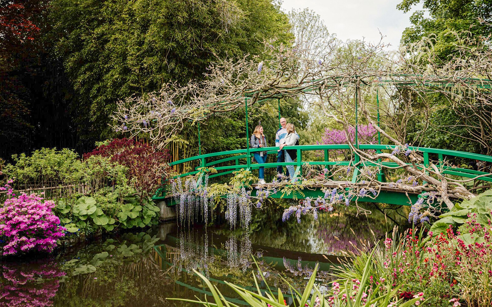 Visitors on Monet's Japanese bridge in Giverny garden, surrounded by lush greenery and flowers.