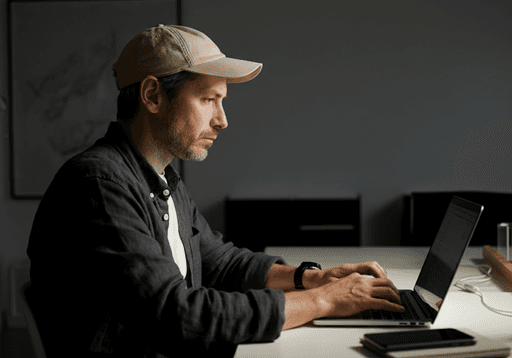 Man sitting at a desk typing on a laptop in a softly lit room, wearing a light-colored cap and casual clothing.