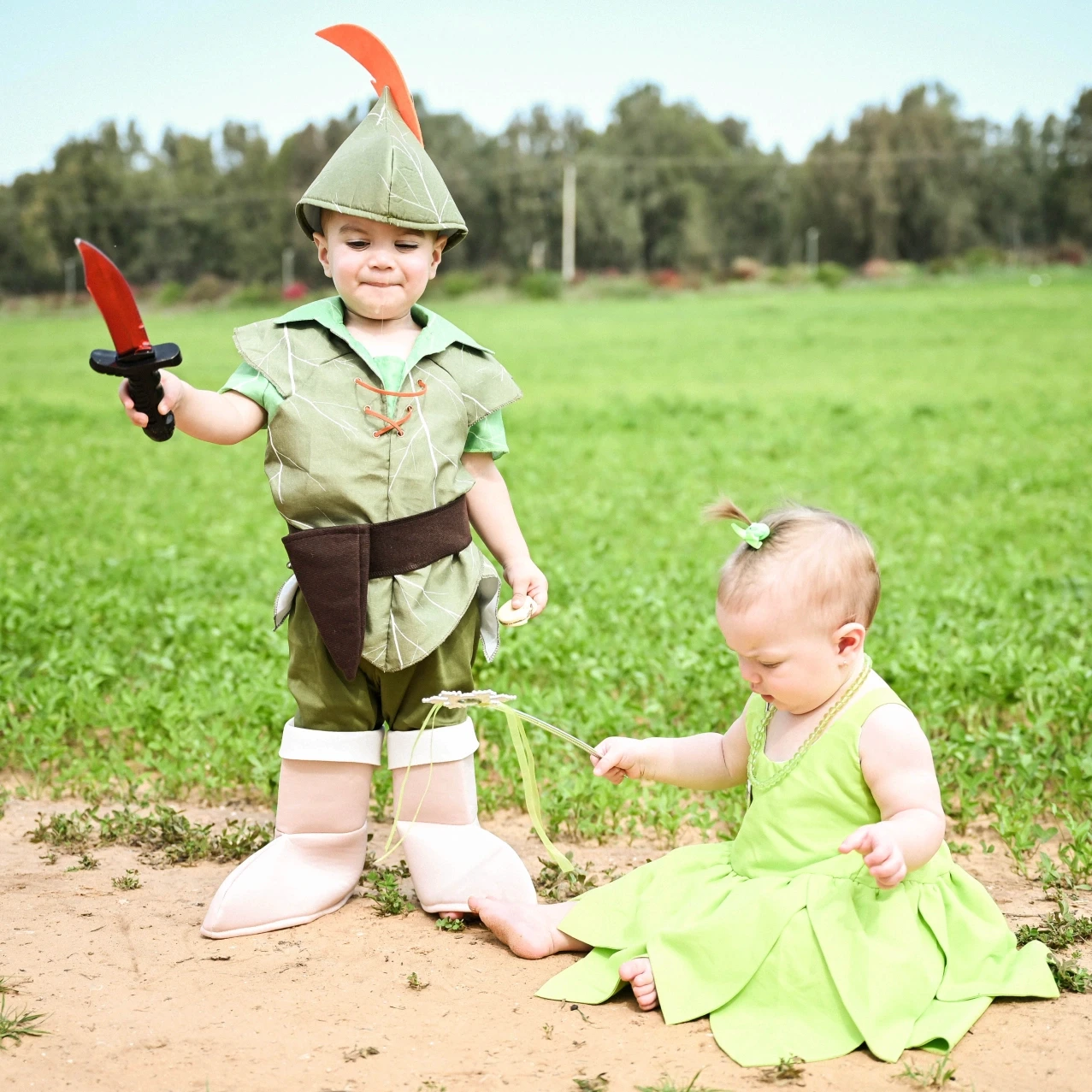 שני ילדים קטנים מחופשים בטבע לרגל חג פורים: פעוט בתחפושת פיטר פן אוחז בחרב צעצוע ותינוקת בתחפושת פיה ירוקה יושבת על הקרקע. צילום ילדים אמנותי באור יום מאת הצלמת שירי עציץ Shiriz Photography