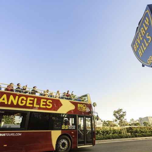 A double-decker tour bus in Los Angeles passes by a sign for Beverly Hills under a clear blue sky, with passengers seated on the top deck.