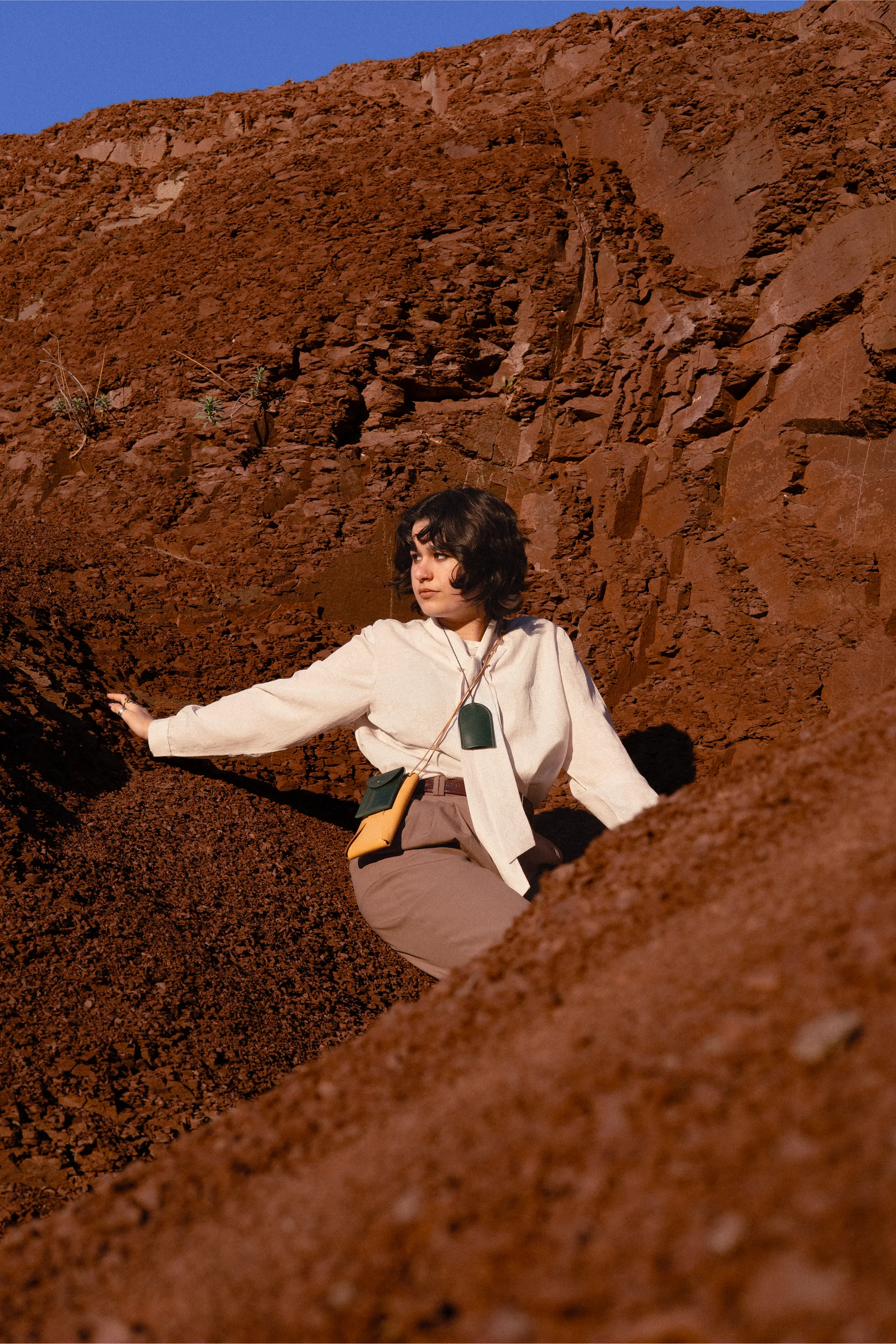 Anaïs Brancheriau wearing a light shirt and taupe trousers, seated between red rock walls, with two hanging leather accessories.