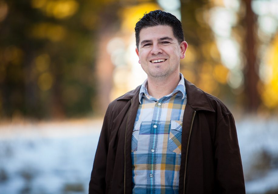 A photo of an attractive man in his late thirties with short dark hair, wearing a blue plaid shirt and a brown jacket, smiling at the camera. The background is a park in winter. The square-shaped app icon logo for the Instagram profile picture has orange tones and a light pink background.