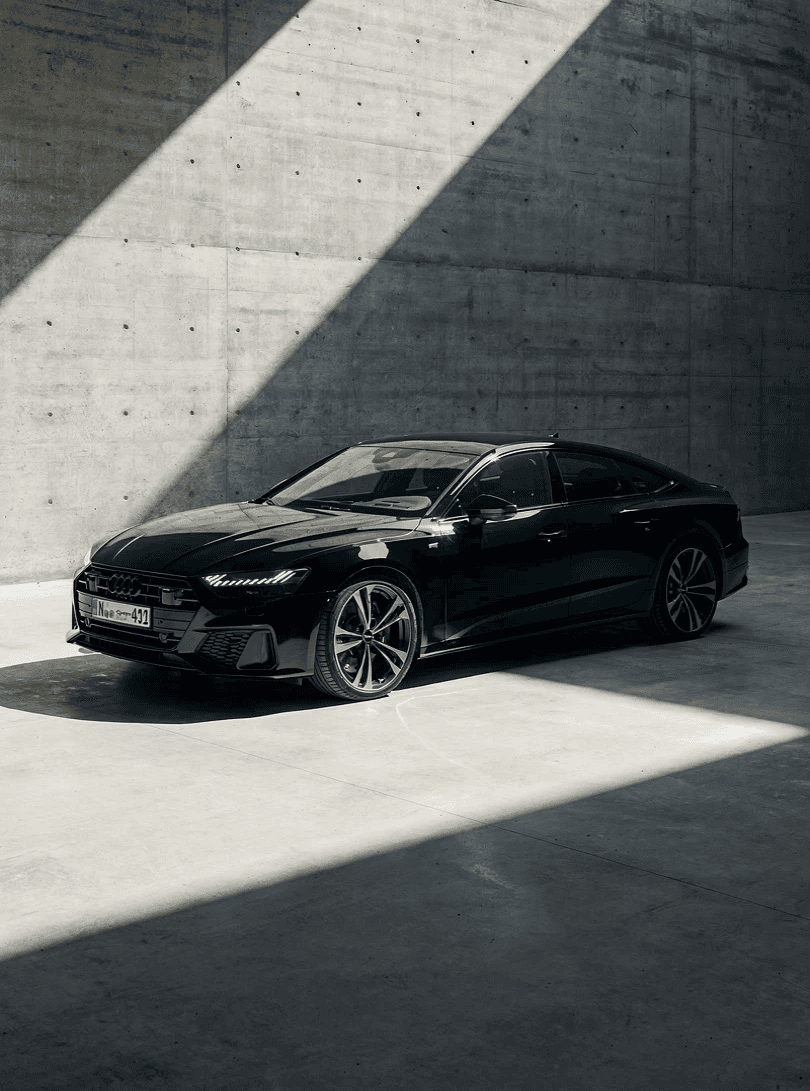 Sleek black car in a modern concrete space, partially lit by a diagonal beam of sunlight, creating a dramatic contrast. The scene feels sophisticated and elegant.