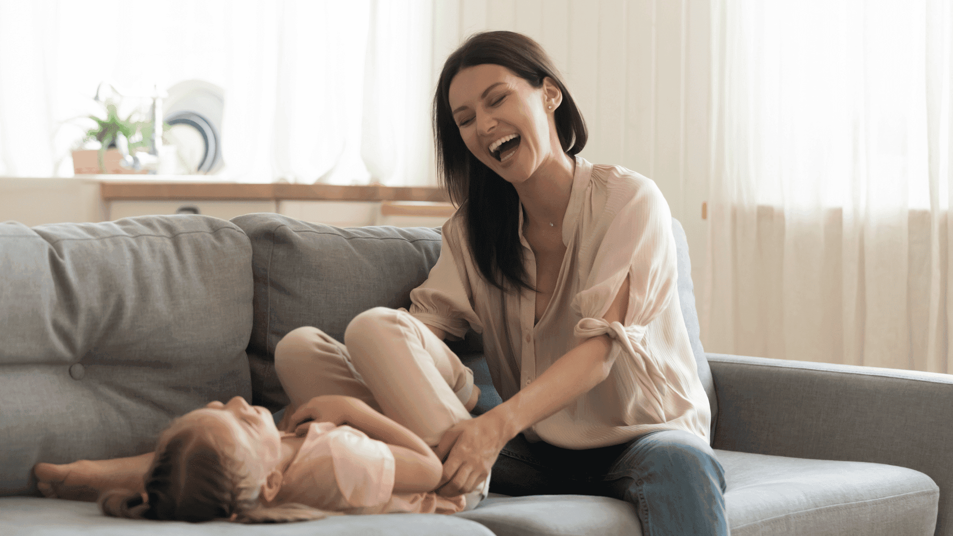 A mother and daughter playing on the sofa, both laughing and looking happy together.