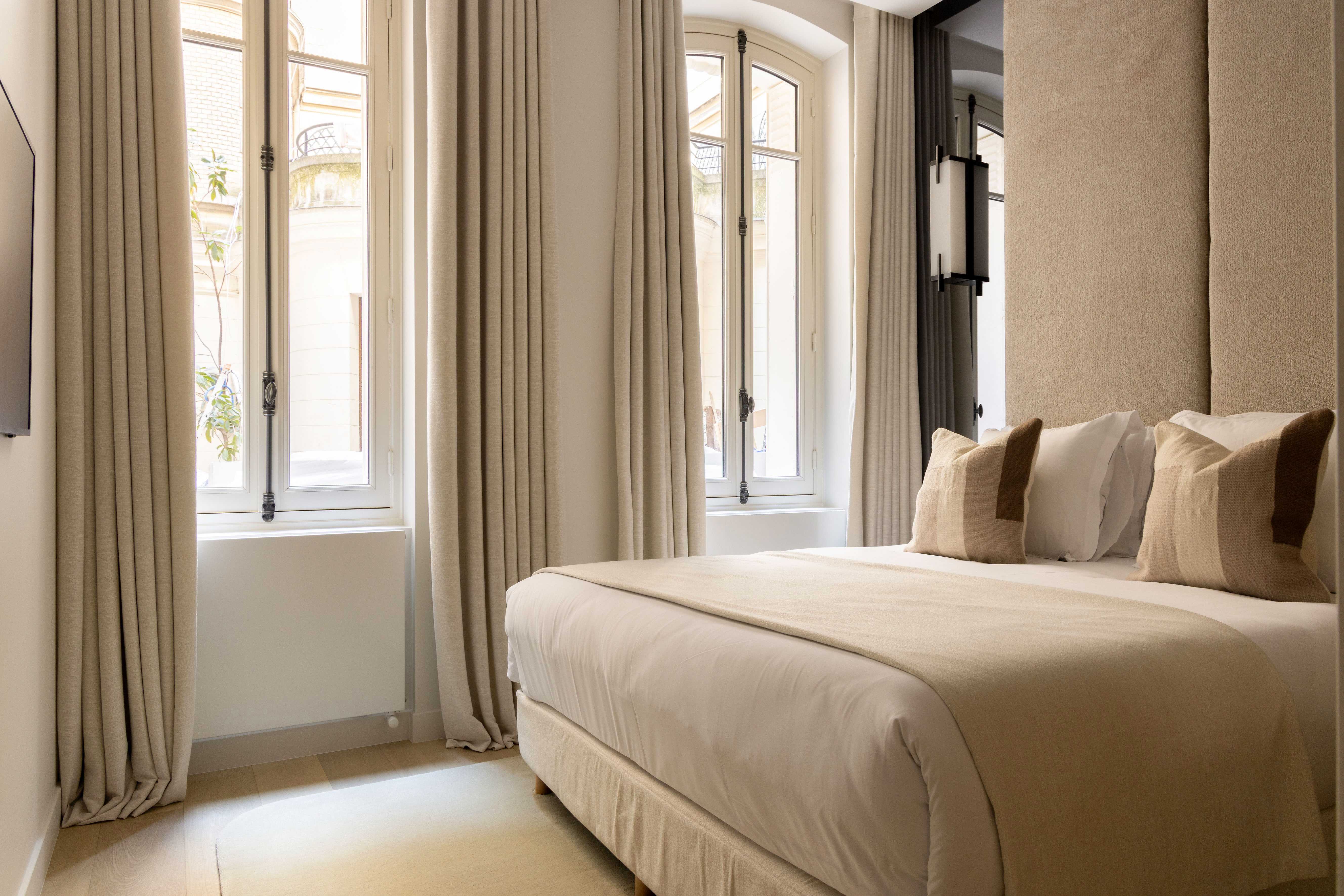 Chambre avec lit double, haute tête de lit rembourrée beige et fenêtres cintrées donnant sur la rue