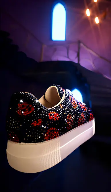 A close-up of a black platform sneaker adorned with shimmering red and orange crystals, set against a moody, purple-lit interior with a glowing blue window