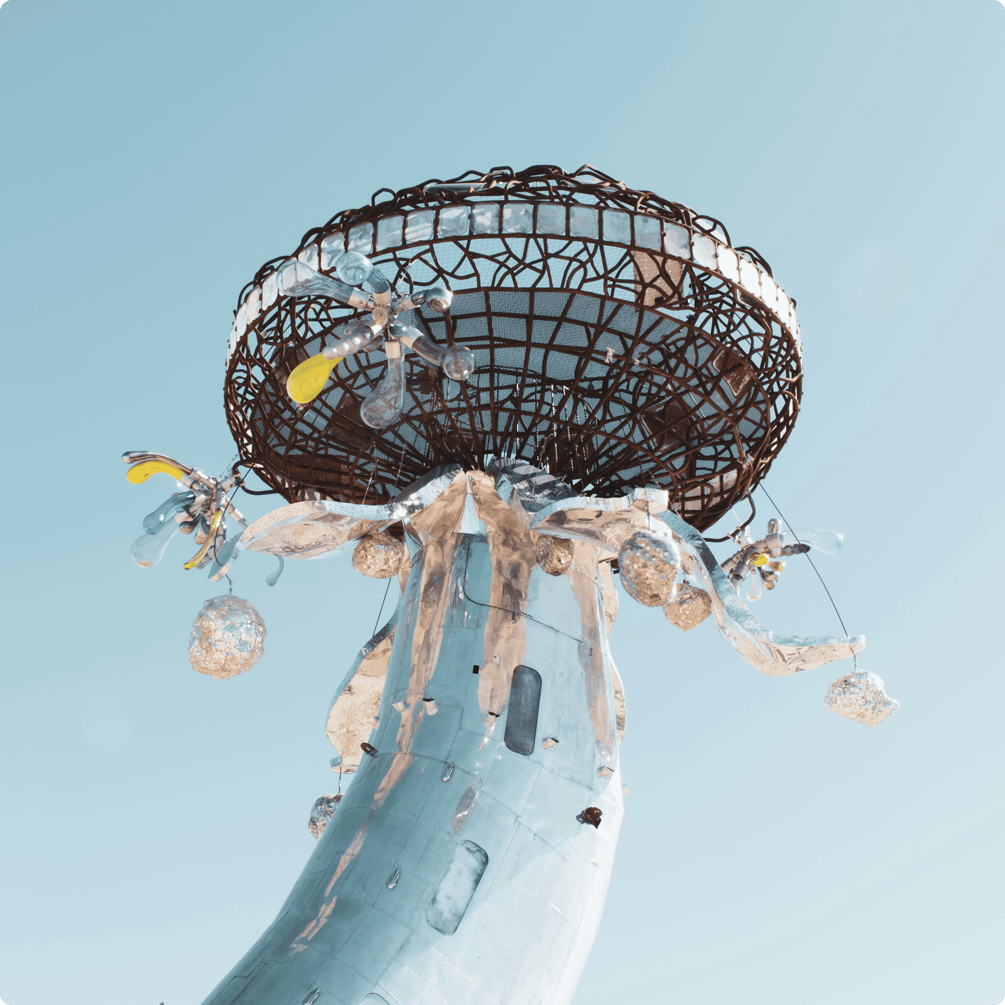 Low angle view of a tall, futuristic silver tower sculpture topped with a round mesh canopy against a clear light blue sky