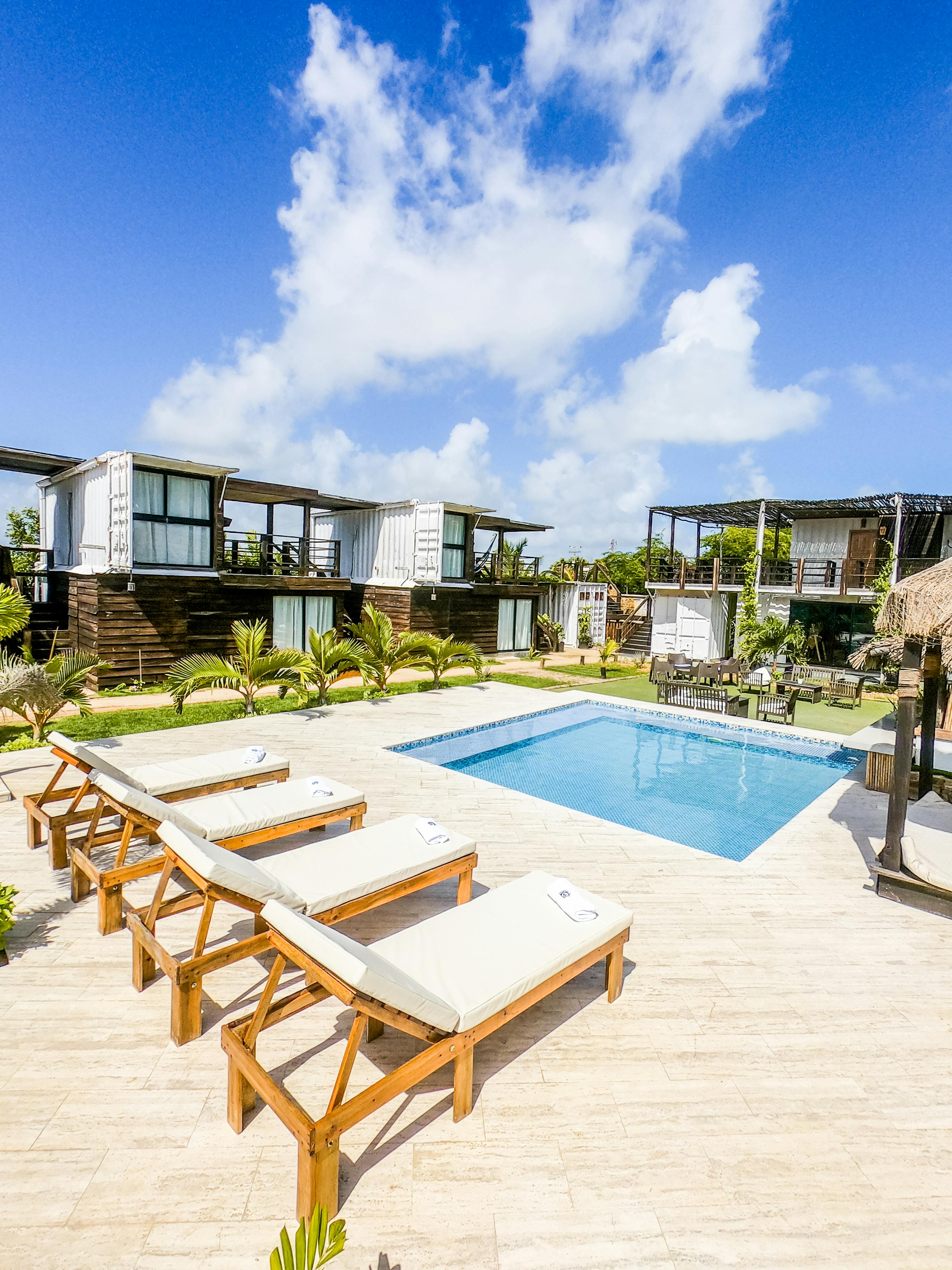 A luxurious outdoor swimming pool area with wooden lounge chairs lined up on a tiled deck, surrounded by modern, minimalist shipping container homes and lush greenery under a bright blue sky.
