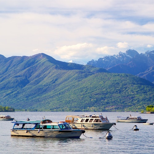 Multiple boats anchored on a calm lake with green, forest-covered mountains and a partly cloudy sky in the background.