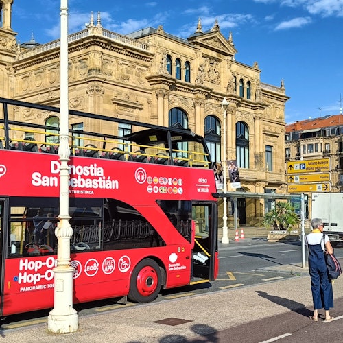 A red double-decker tour bus in San Sebastián stops on a city street with historic buildings and a hill in the background.