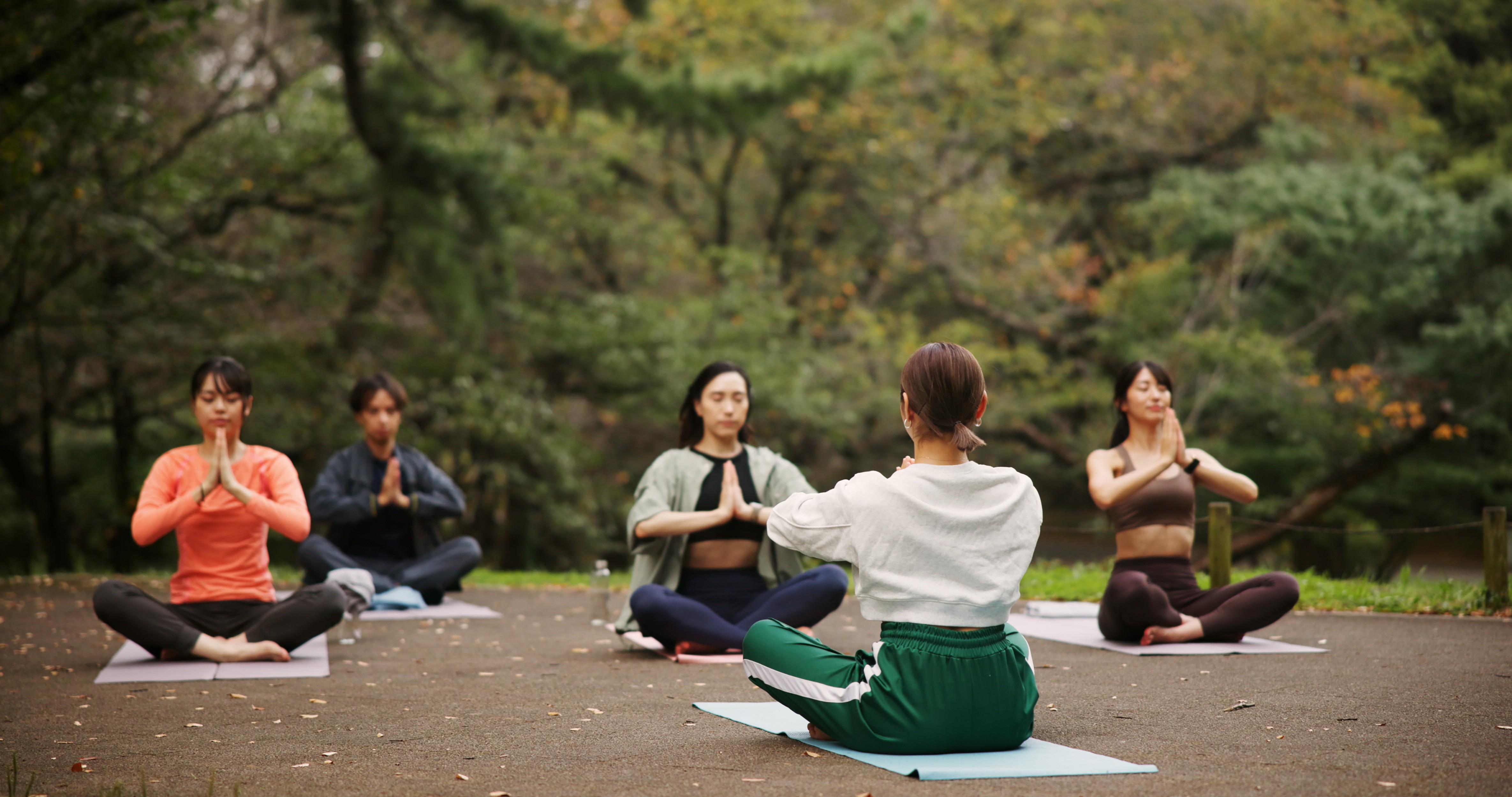 A group of people sitting cross-legged on mats in a park holding their hands in a prayer pose during an outdoor yoga or meditation session.