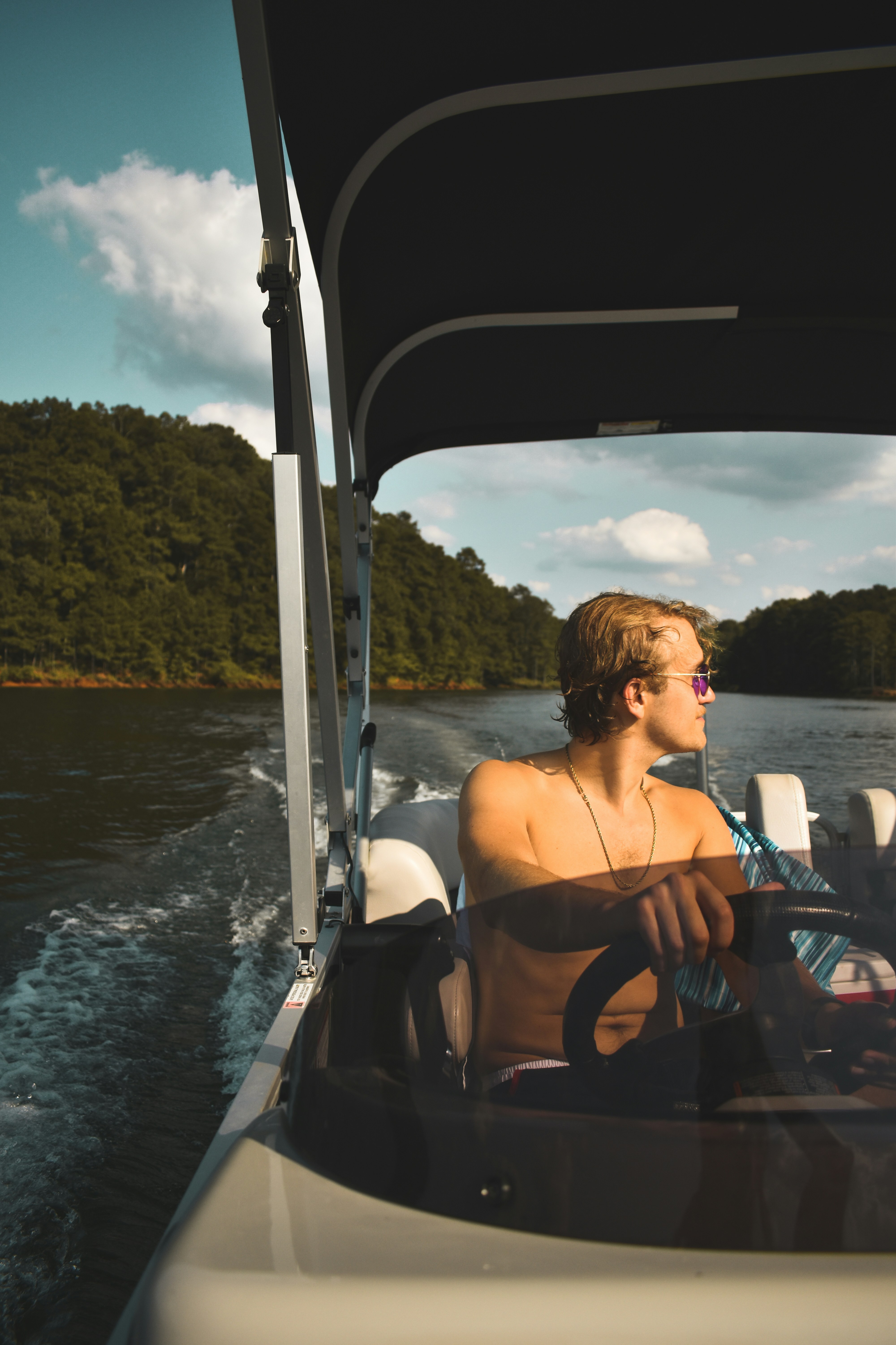 A person wearing sunglasses navigates a motorboat on a tranquil lake surrounded by lush, green trees under a partly cloudy sky.