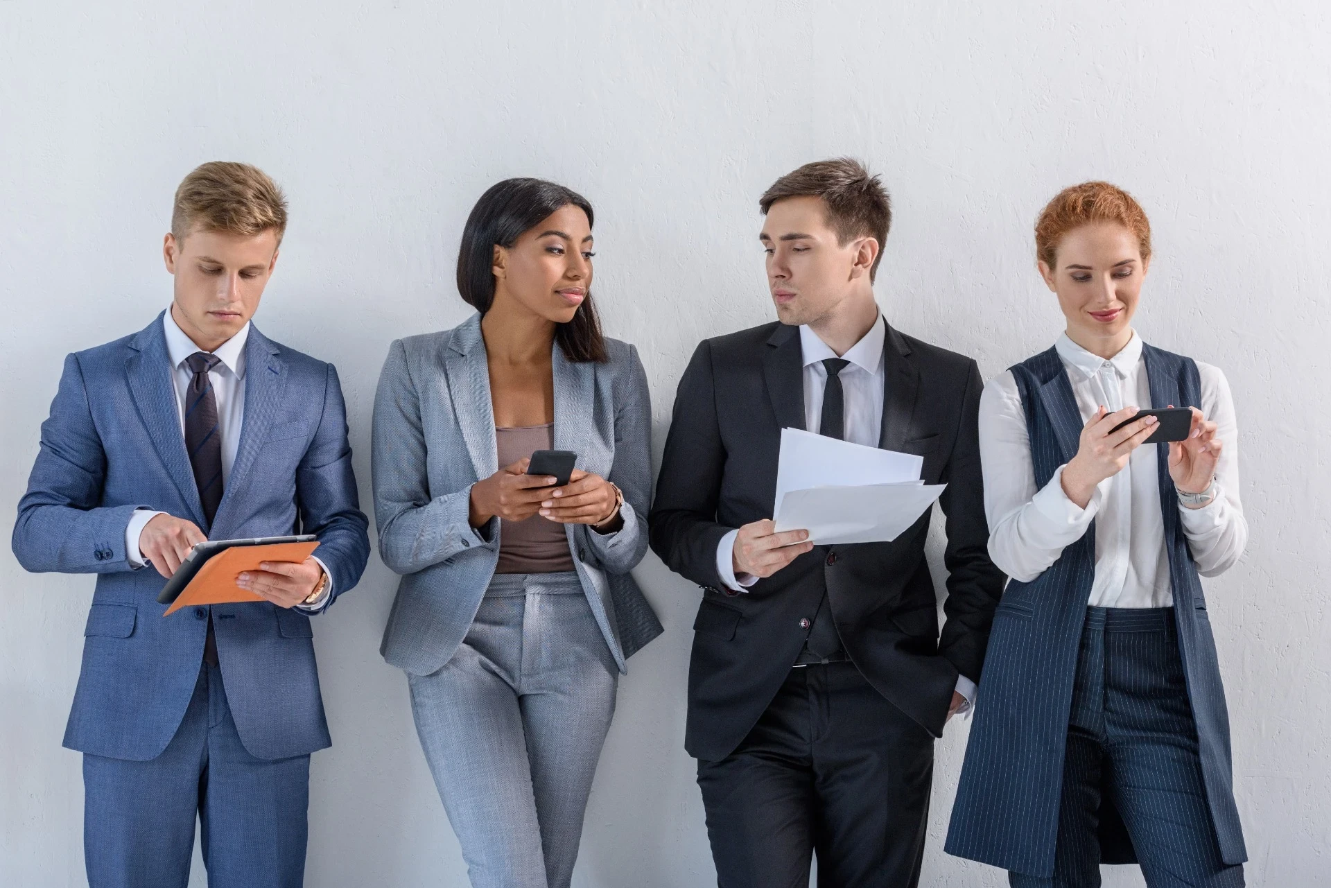 A imagem mostra quatro jovens profissionais em um ambiente formal, provavelmente durante uma pausa ou em um momento descontraído antes de uma reunião. Eles estão vestidos em trajes de negócios e parecem engajados em diferentes atividades, como revisar documentos, usar dispositivos móveis e um tablet. A postura de cada um indica concentração, com um leve toque de interação entre eles. A composição da imagem, com os profissionais alinhados contra uma parede branca, transmite um ambiente corporativo moderno e dinâmico.