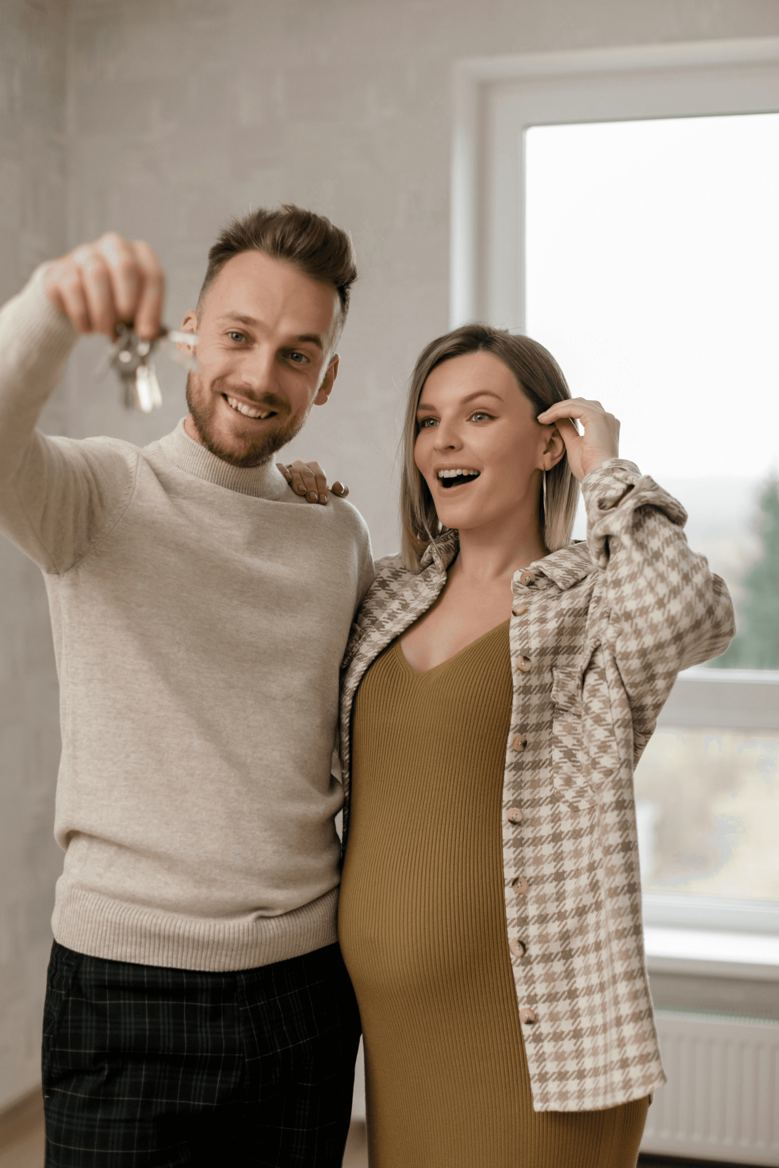 A happy couple, one holding keys and the other with a hand on their baby bump, smiling in a bright room.
