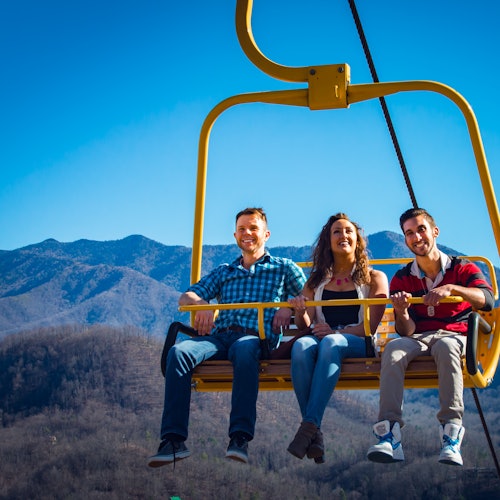 שלושה אנשים מחייכים ורוכבים על מעלית סקי צהובה עם נוף הרים ברקע תחת שמיים כחולים וצלולים.