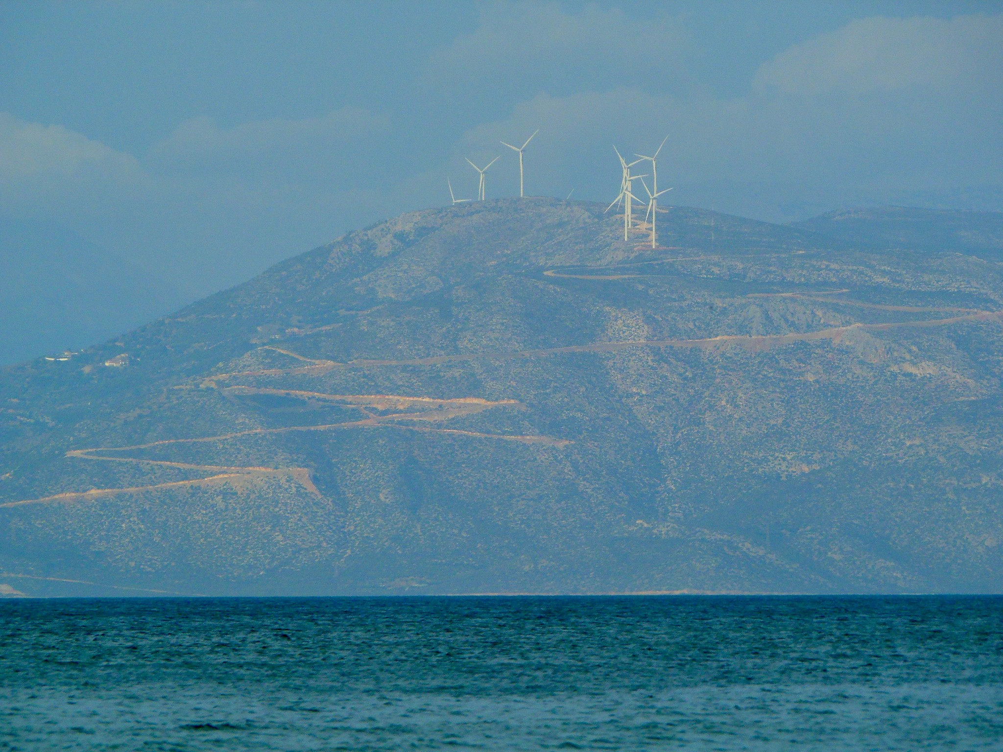 Eine Aussicht auf einen Berg mit mehreren Windturbinen auf seinem Gipfel, vor einem dunstigen Himmel. Im Vordergrund ist der ruhige Ozean zu sehen, und die Landschaft zeichnet sich durch raues Gelände aus, auf dem einige kurvenreiche Straßen entlang des Berges sichtbar sind.