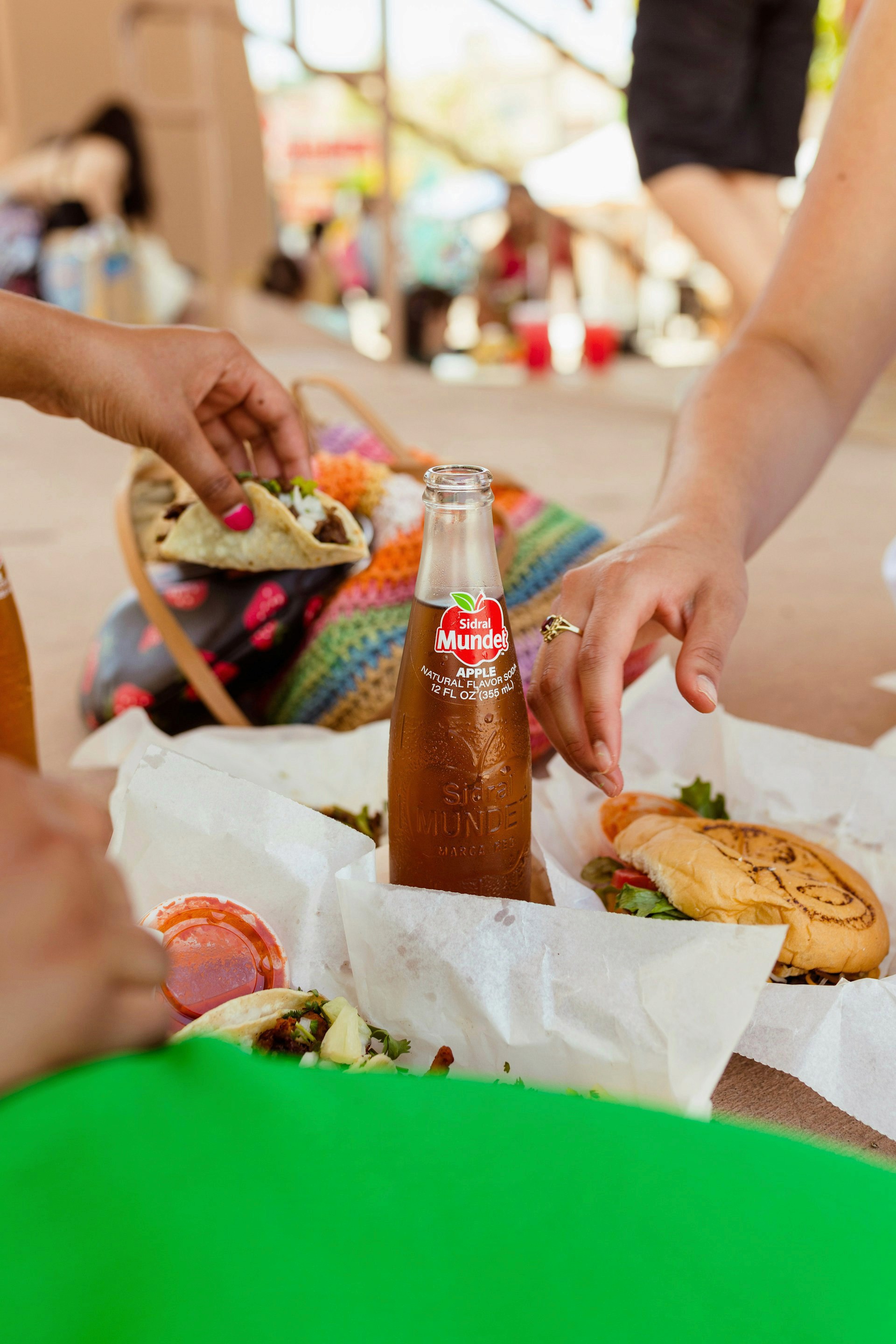 People's hands reaching for street tacos and a sandwich wrapped in white paper, centered around a cold bottle of apple soda.