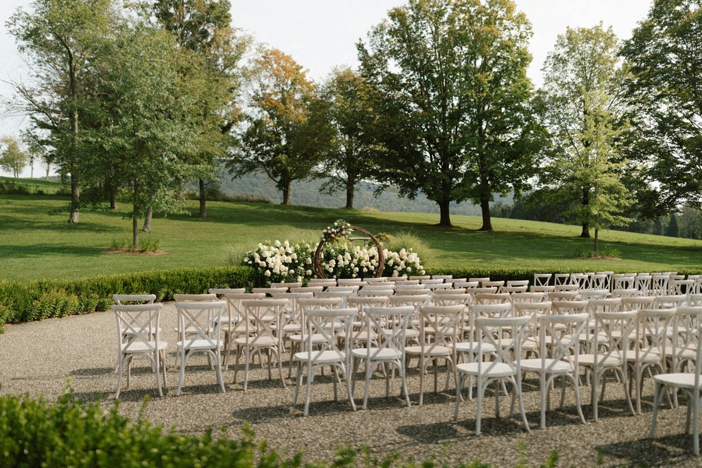 Outdoor ceremony seating with white crossback chairs facing a circular floral arch surrounded by white hydrangeas on a manicured estate lawn