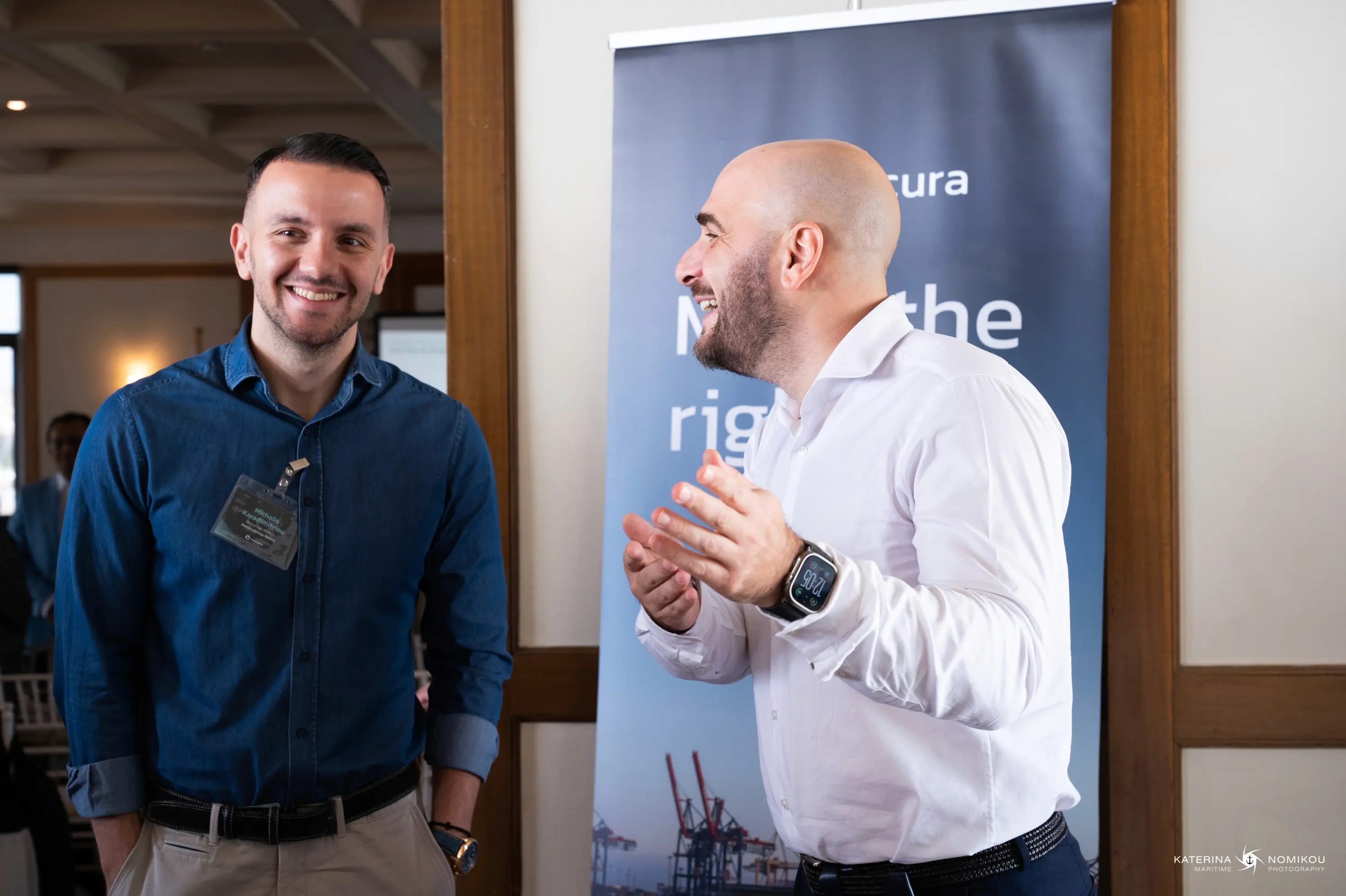 Two men are smiling and talking together in a professional setting, with a banner in the background.