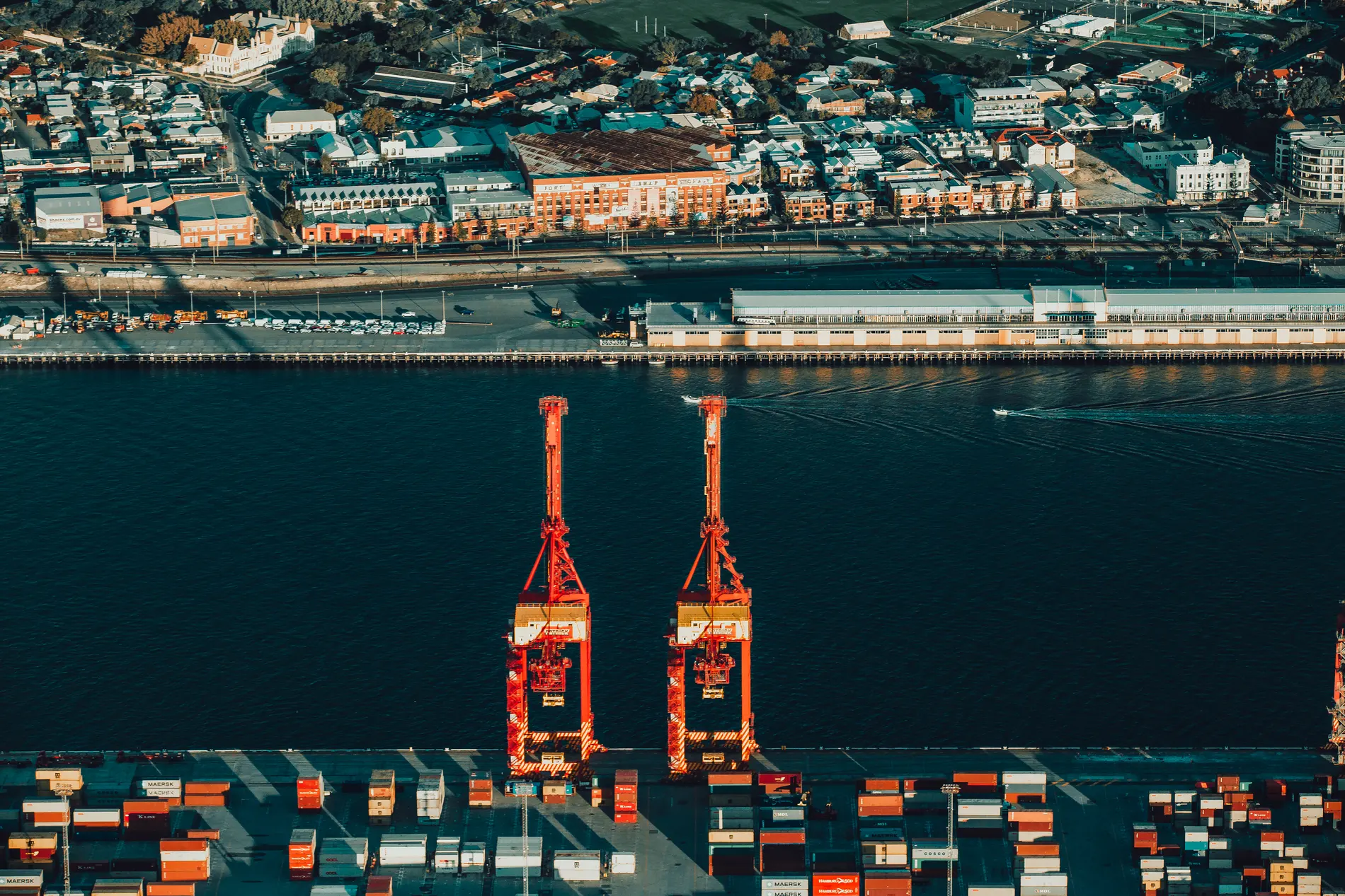 Aerial of two red harbour cranes and container stacks beside long heritage port sheds