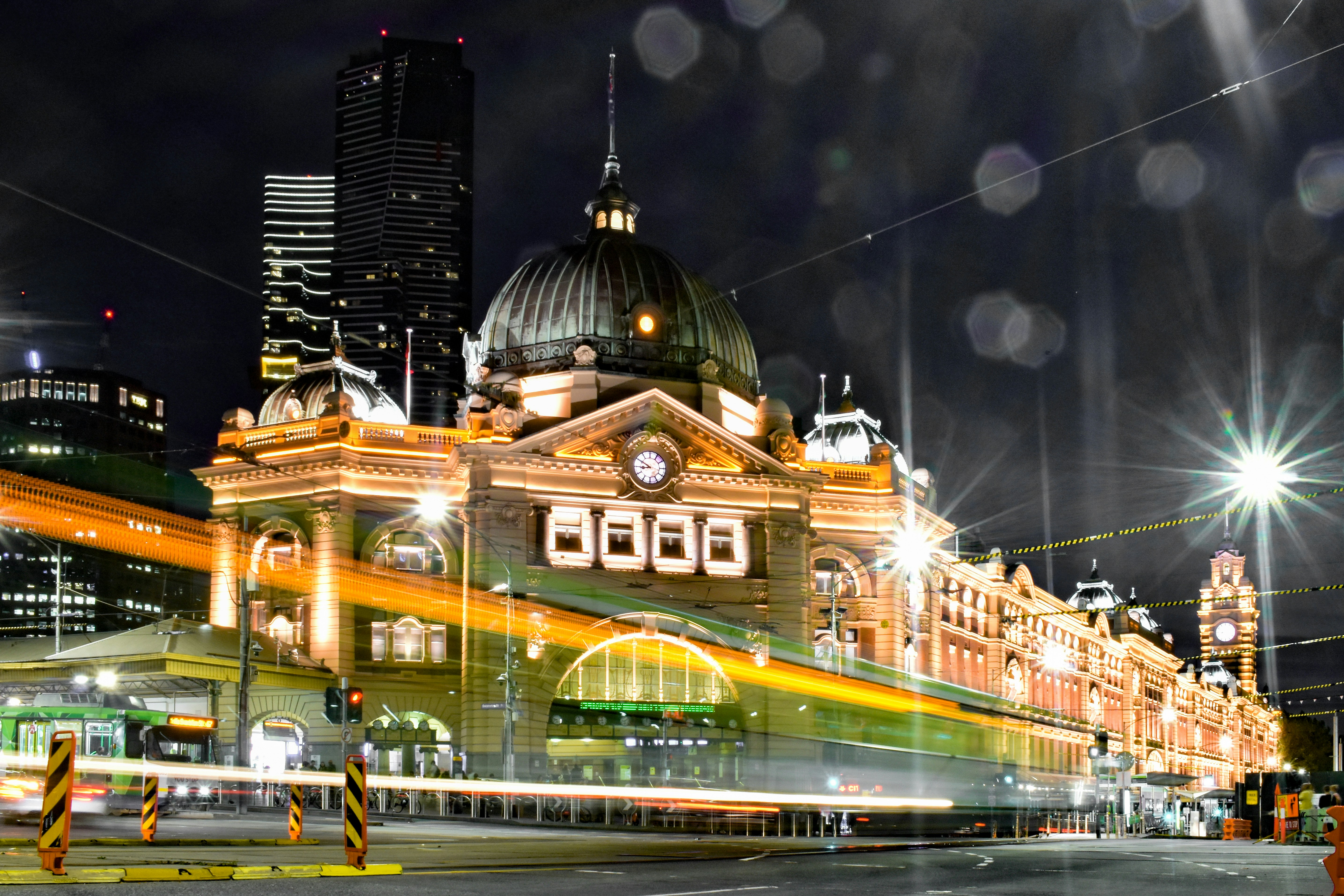 A large building, namely Flinders Street Station in Melbourne, at night with skyscapers in the background.