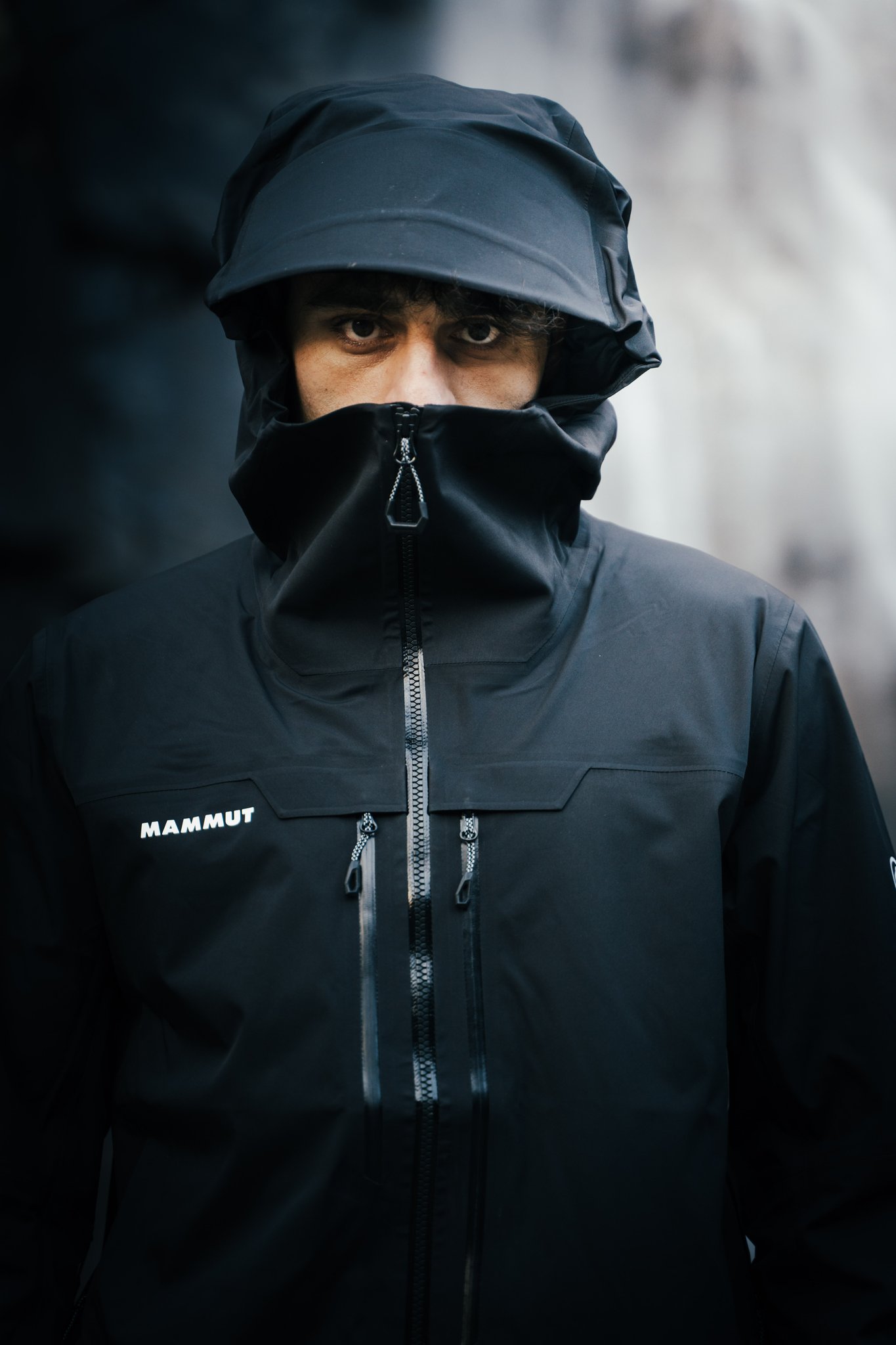 A person wearing a black hooded jacket, partially obscured face, stands against a dark backdrop.