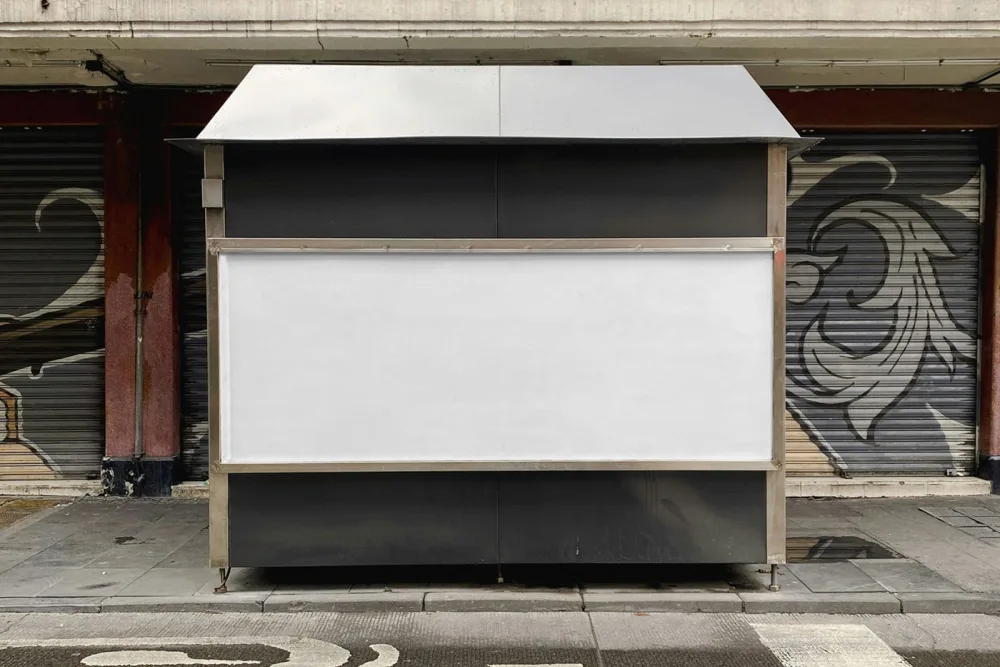 Mockup of an outdoor banner displayed on a metal structure in an urban environment. The design features bold black text and geometric yellow elements, set against a background of graffiti and closed storefronts for a gritty, modern vibe.