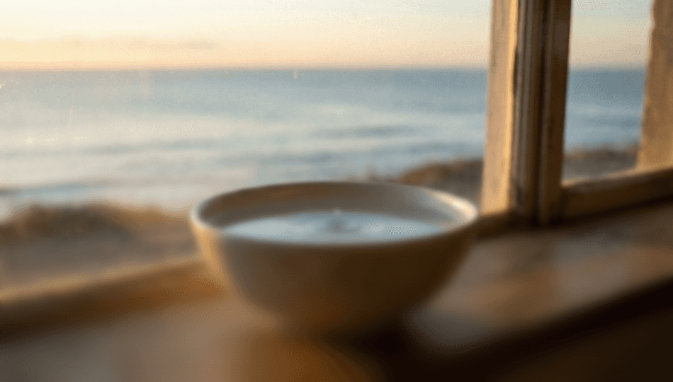A photograph of a simple, elegant ceramic bowl filled with clear water, sitting on a wooden windowsill