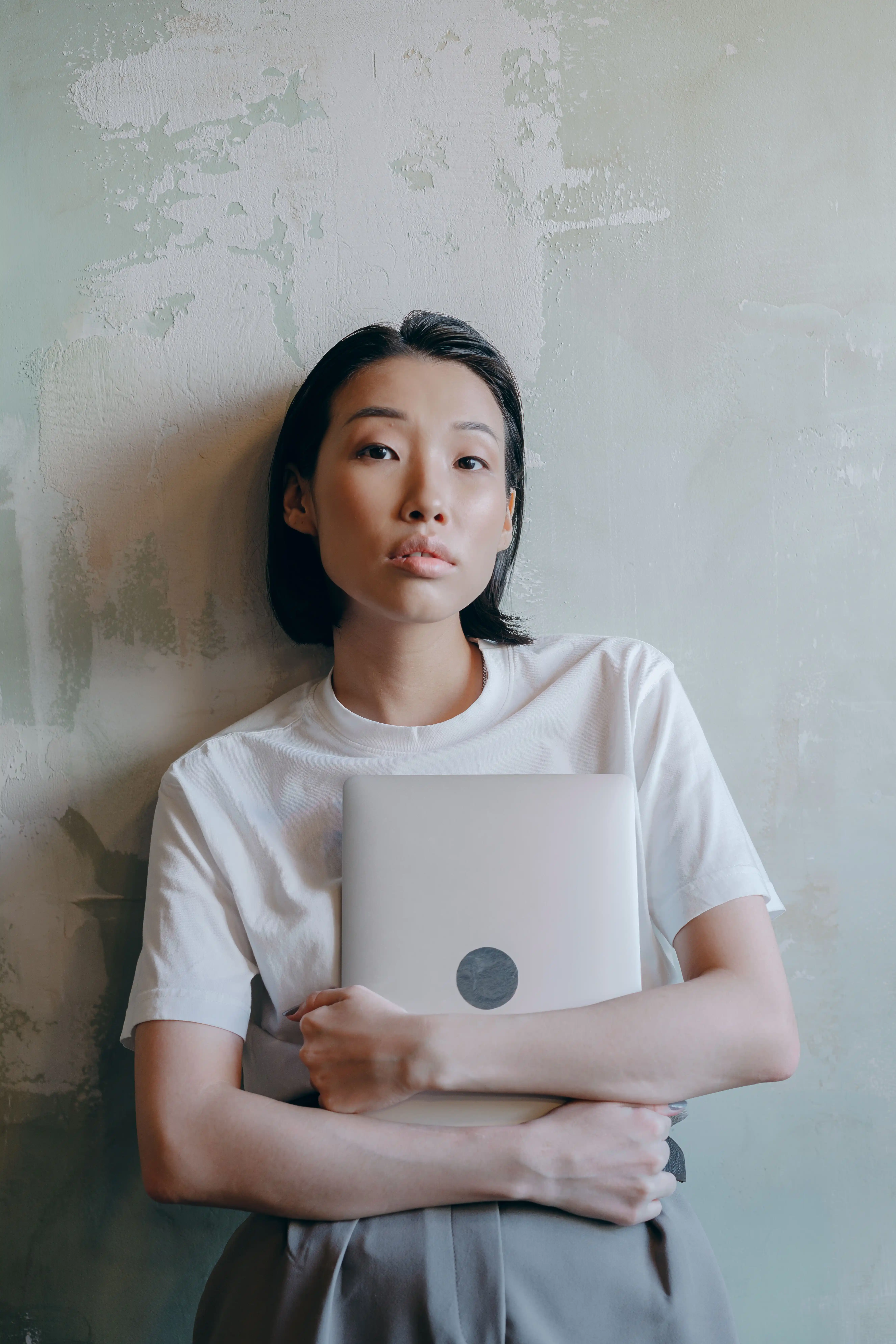 A young woman with short dark hair wearing a white t-shirt, holding a closed laptop against her chest while leaning against a textured, light green distressed wall.