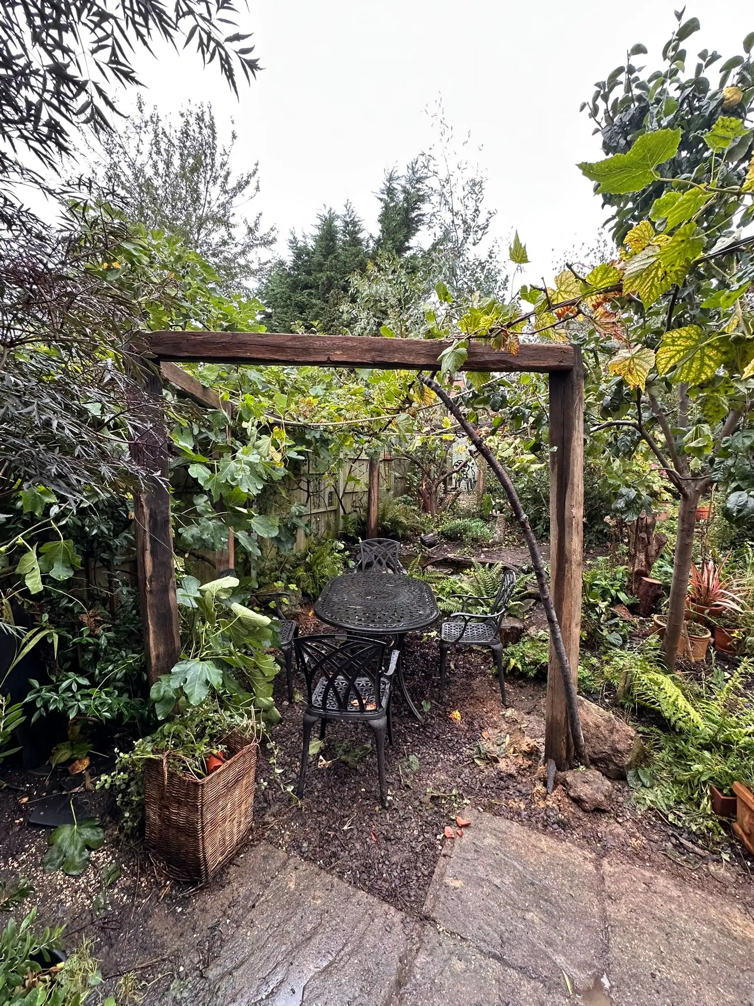 A lush garden entrance with wooden arch, surrounded by greenery and flowers, and a stone path leading through.